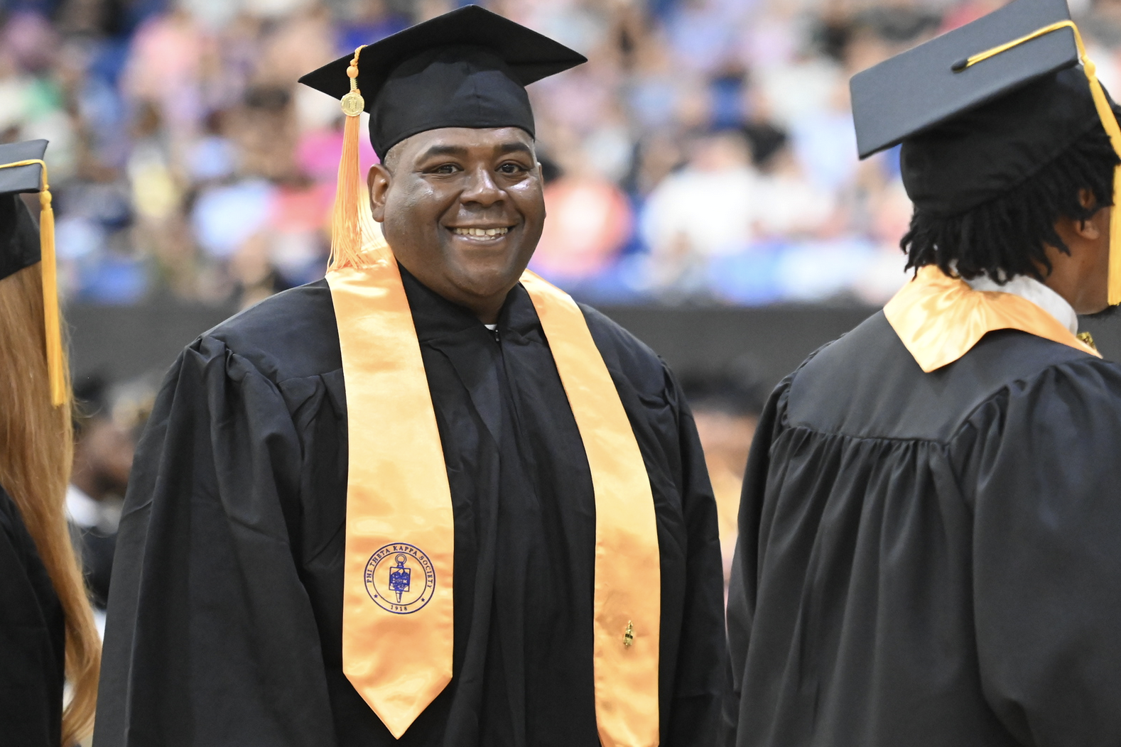 A graduate smiling while wearing a black cap and gown, with a gold stole draped around his neck during a graduation ceremony.