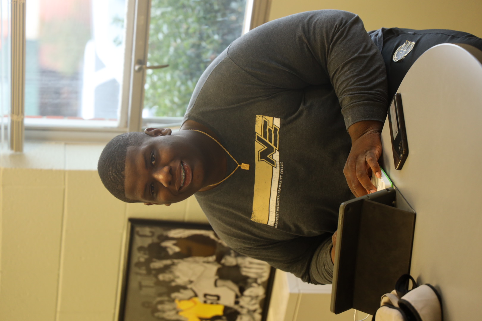 student sitting at a table using an iPad; he is looking at the camera and smiling