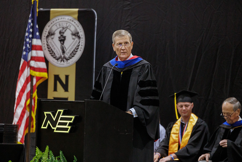 Commencement speaker in academic regalia stands at a podium on a graduation stage, with a U.S. flag and seated graduates in the background.
