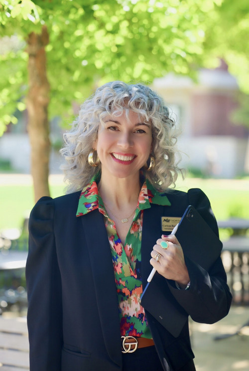 Female with short silver curly hair wearing a black floral blouse.  She is looking at the camera, smiling, with her arms crossed.
