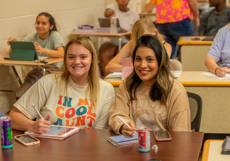 Two female students sitting in desks in a classroom with their iPad.
