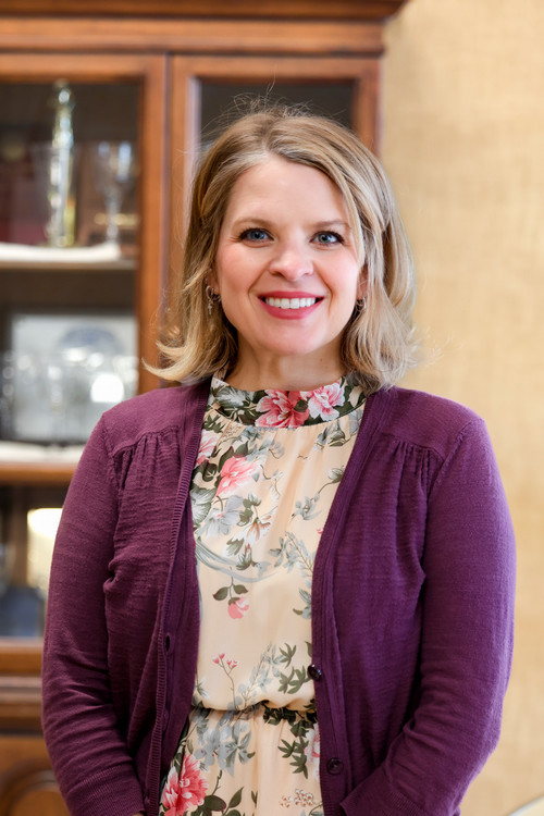 Blonde female in a classroom setting smiling at camera and wearing a floral top and purple cardigan.