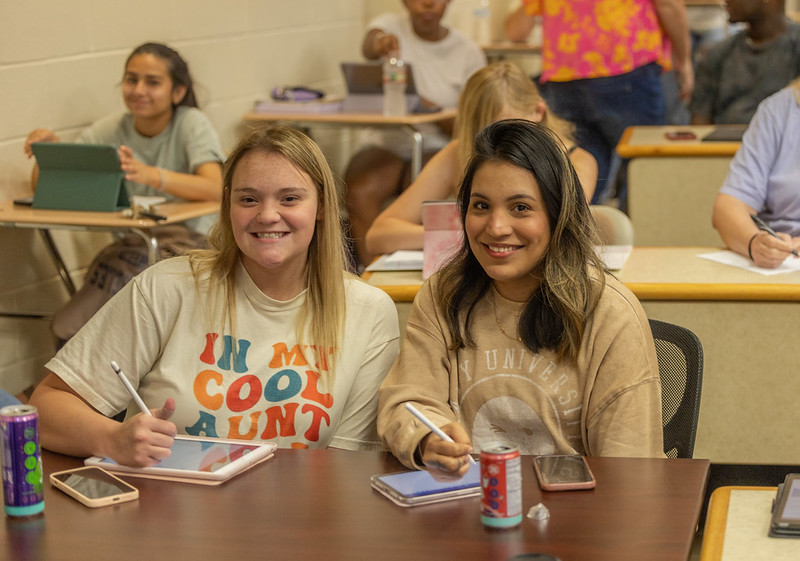 two female students sitting in a classroom smiling at the camera