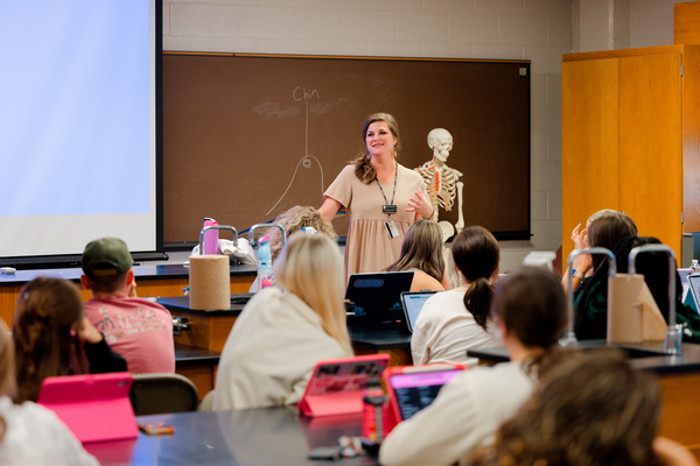 Teacher at the front of a classroom.  Students are sitting at tables with their iPad.